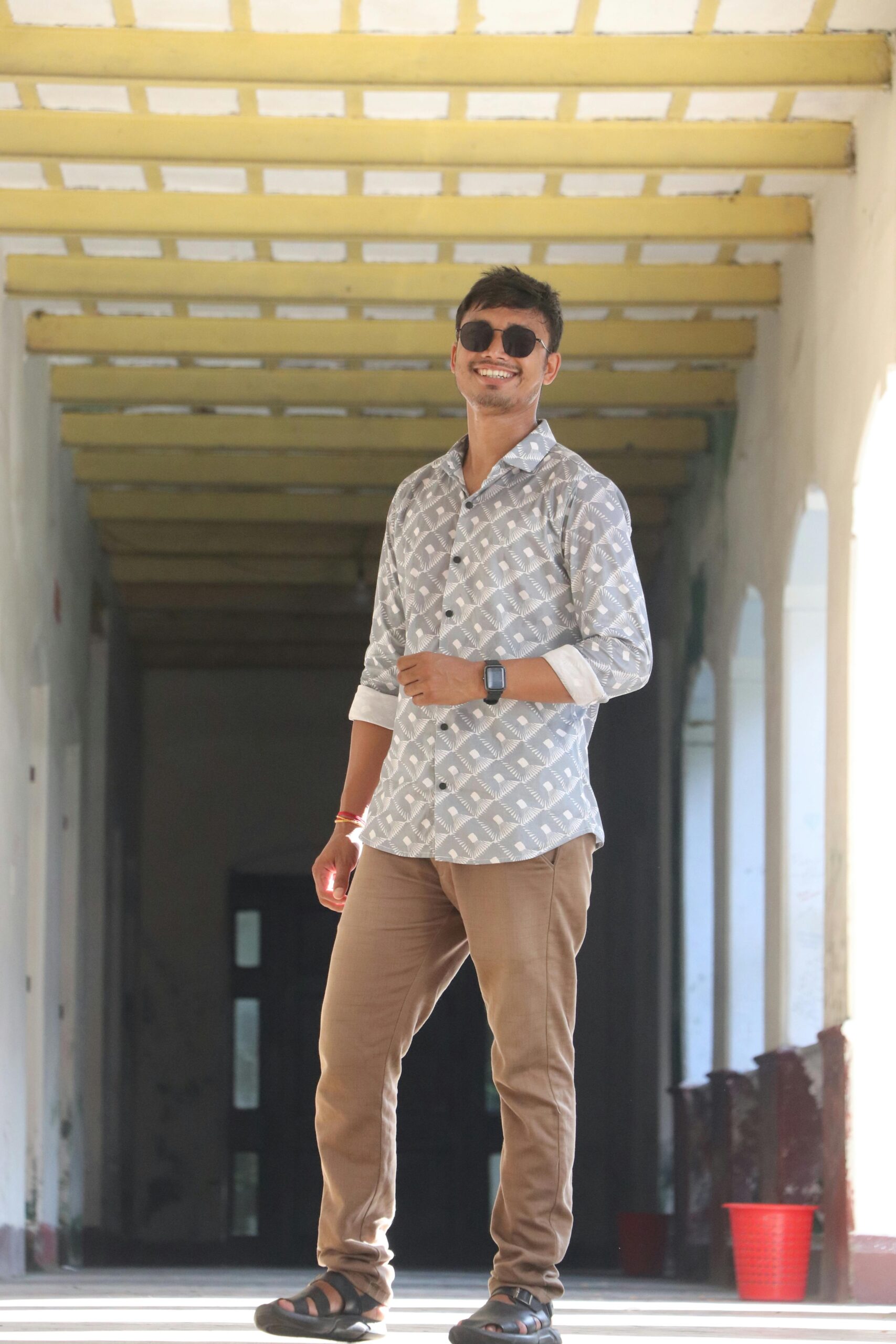 Portrait of a young man smiling in a well-lit corridor, wearing casual attire and sunglasses.