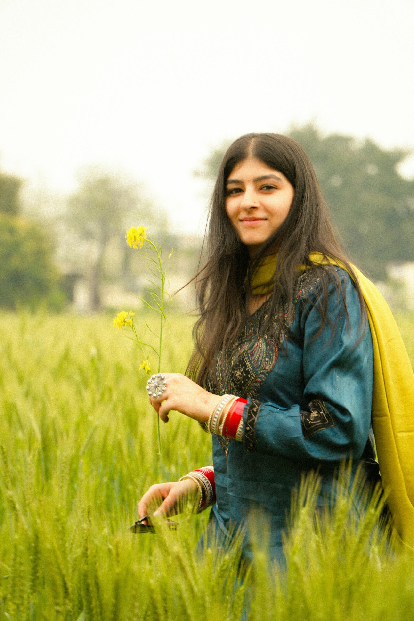 A smiling woman in traditional attire holding a flower in a lush green field.